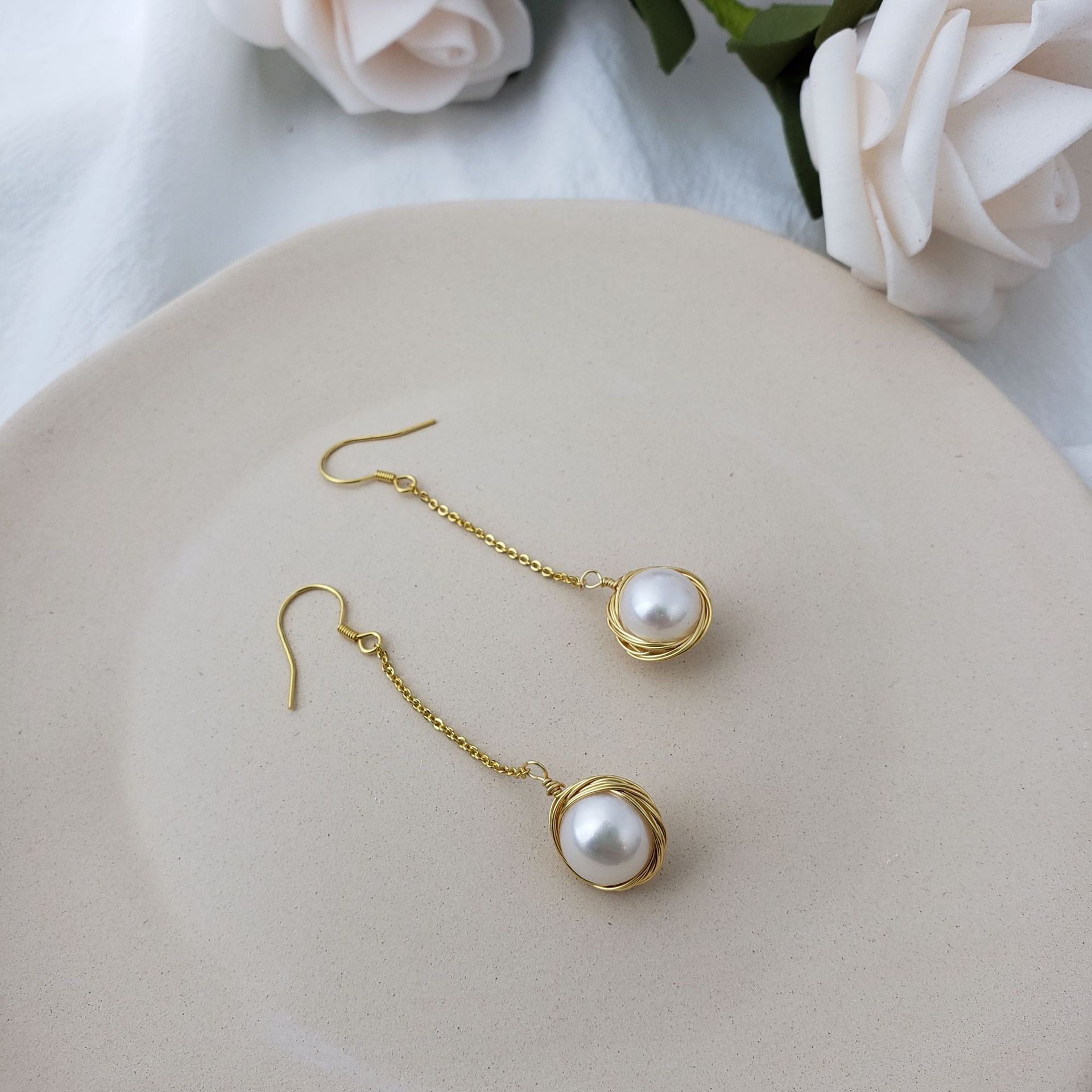 Pair of pearl earrings with gold chains on a light surface with flowers in the background