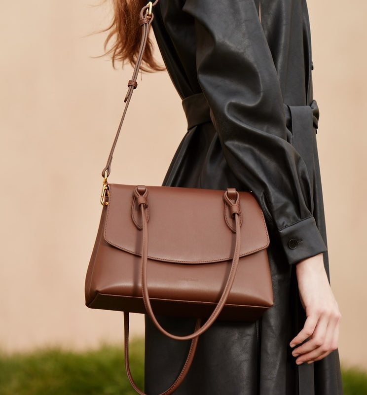Brown leather handbag held by a person wearing a black coat against a beige background