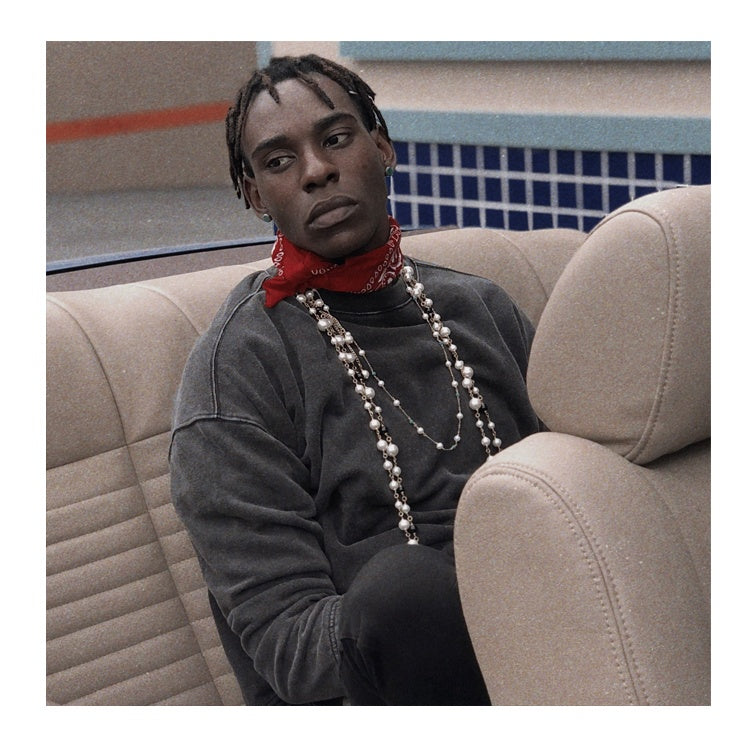 Person sitting on a beige couch wearing a red bandana and beads, with a tiled wall in the background.