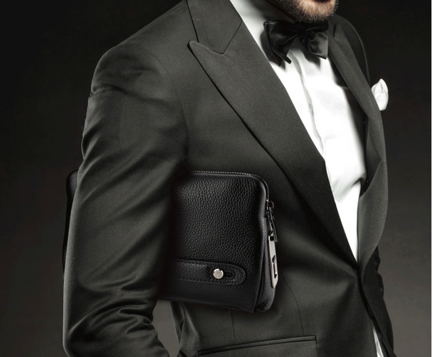Man in a black tuxedo holding a black leather clutch bag against a dark background