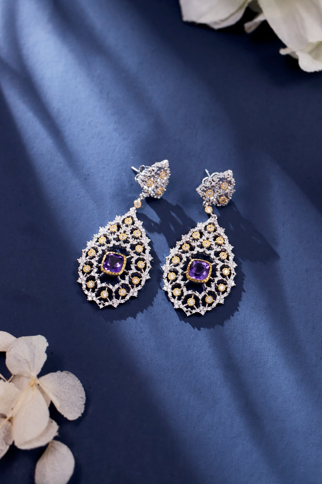 Pair of ornate earrings with purple gemstones on a blue fabric background.