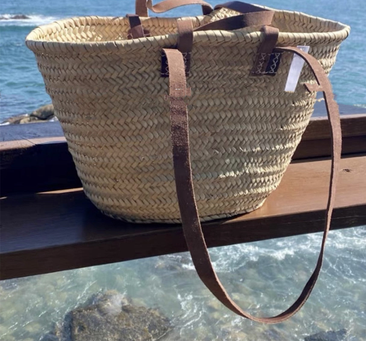 Woven straw bag with leather straps on a wooden ledge by the sea.