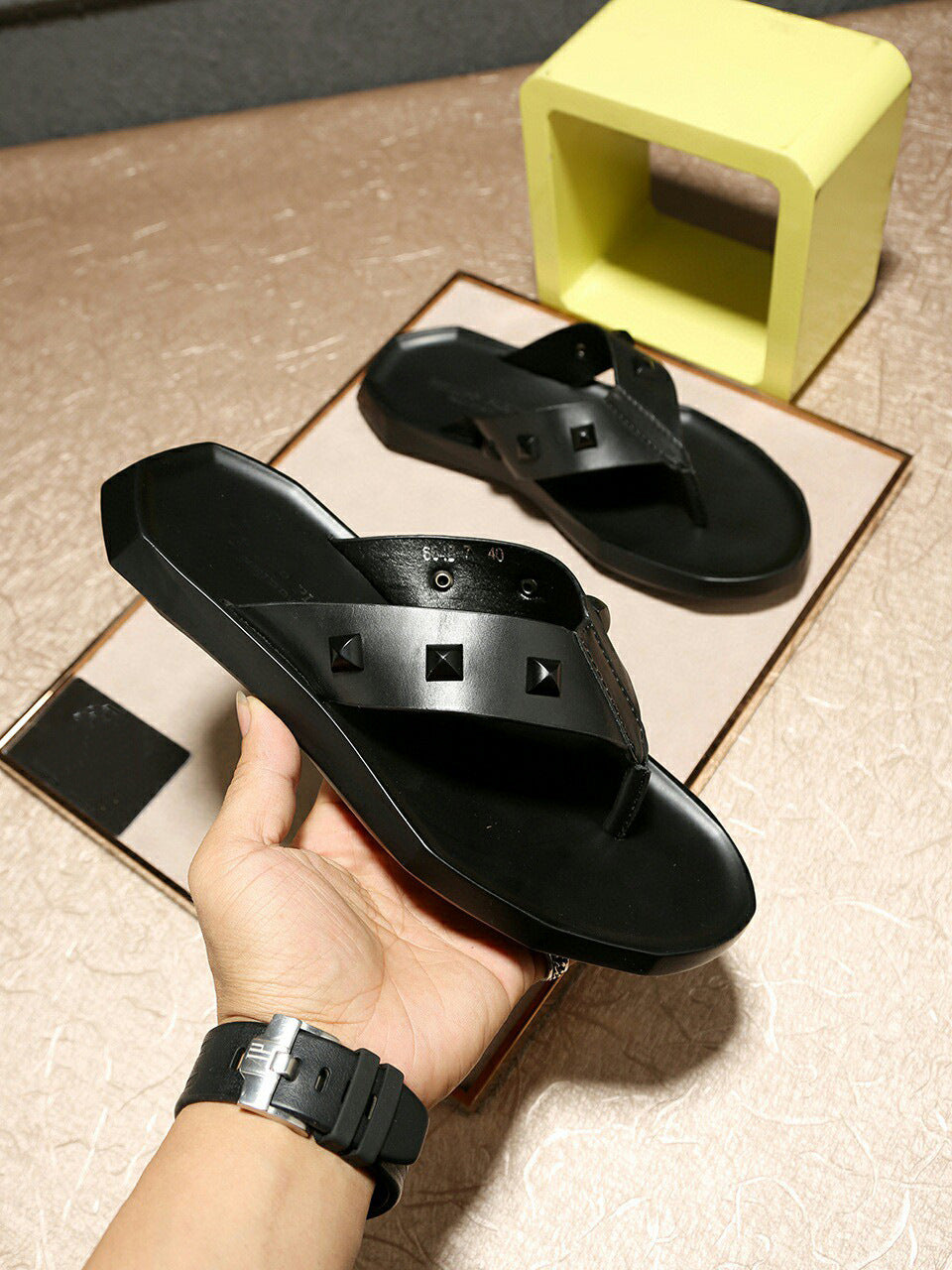 Black sandals held by a hand on a beige surface with a yellow block in the background