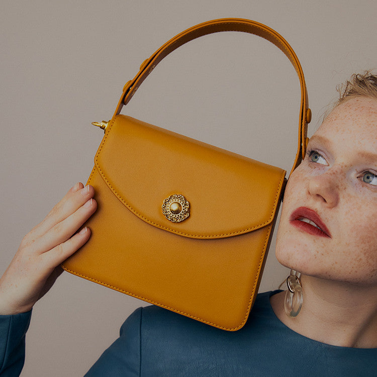 Woman holding a mustard yellow handbag with a decorative clasp against a neutral background