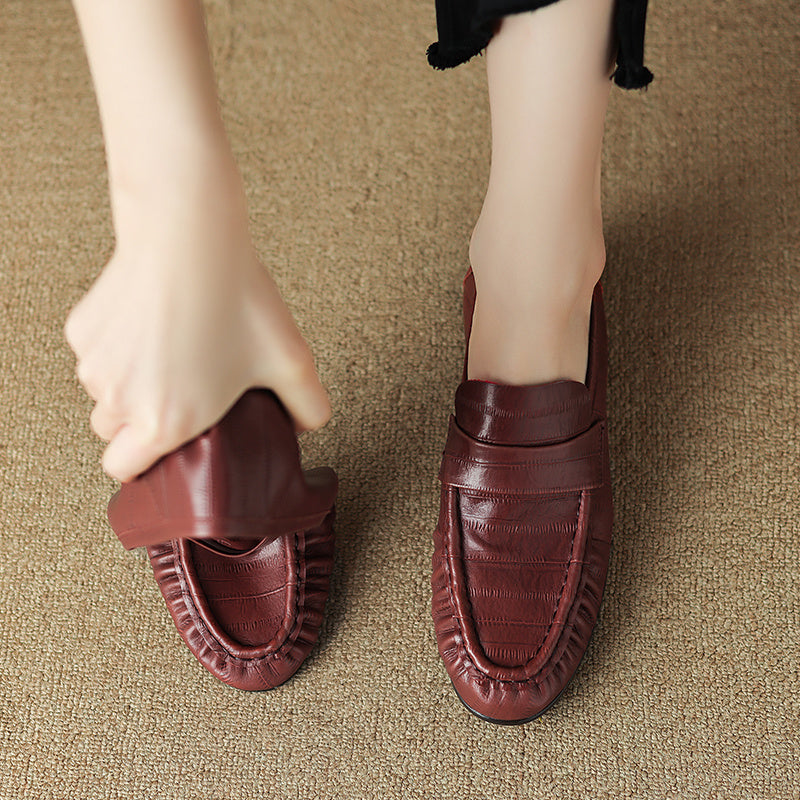 Brown leather loafers on a textured surface with a hand reaching towards one of them.
