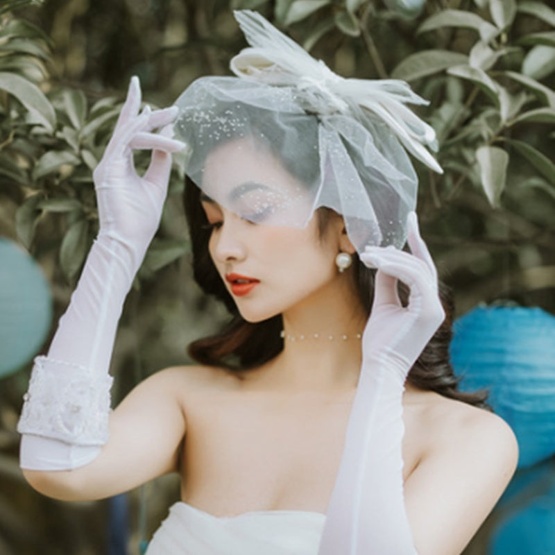 Woman in a bridal outfit with a veil and gloves, standing against a natural background.