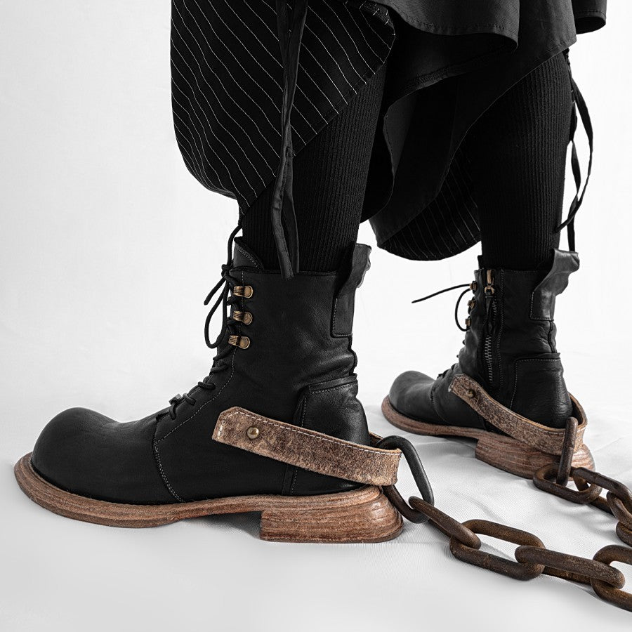 Black leather boots with wooden soles and chains, worn with black pants on a white background
