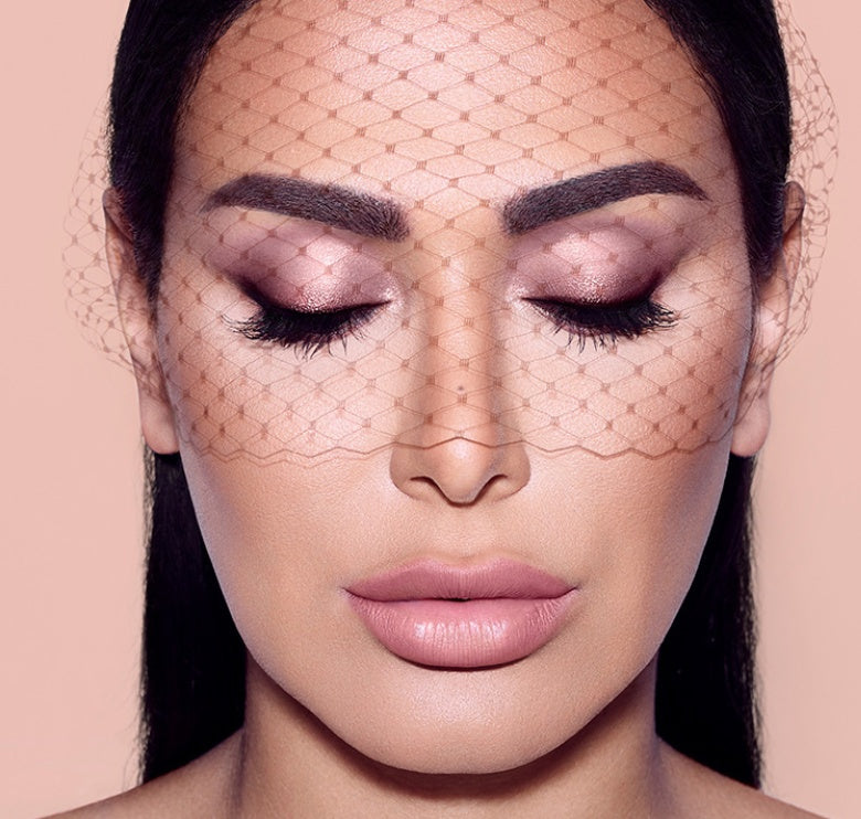 Close-up of a woman's face with makeup, wearing a fishnet veil against a pink background