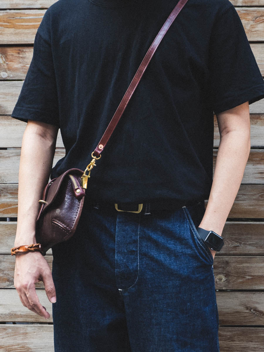 Person wearing a black t-shirt and blue jeans with a brown leather bag over one shoulder against a wooden background.