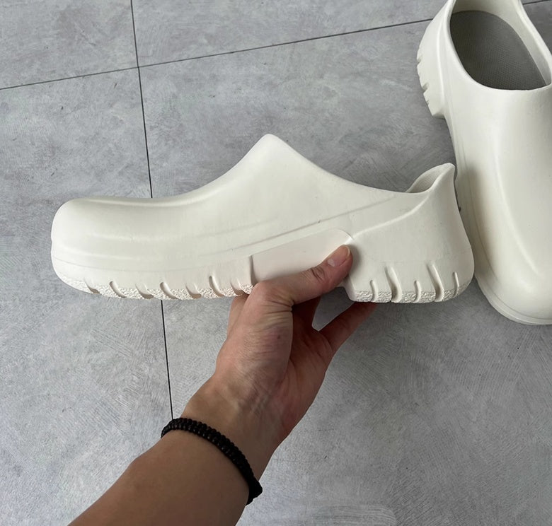 White clogs being held by a hand on a gray tiled floor