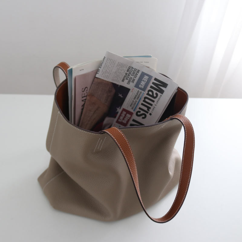 Beige handbag with brown leather straps containing newspapers on a white surface.