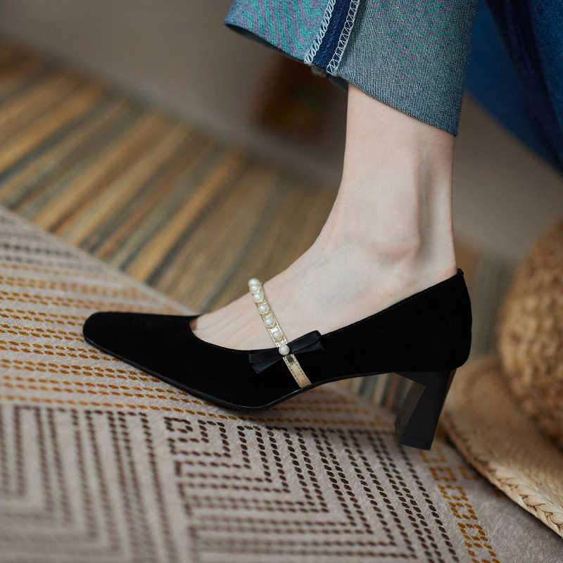 Black suede shoes with pearl bow detail worn by a person on a patterned surface.