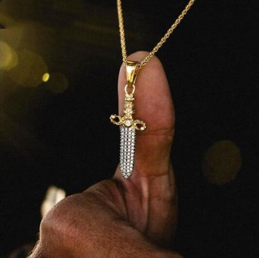 Gold sword-shaped pendant with clear stones on a chain held by a hand against a dark background