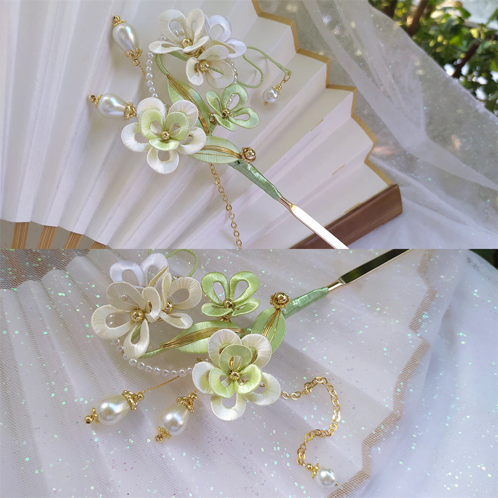 Decorative floral hairpin with green and white flowers and gold accents on a white fabric background.