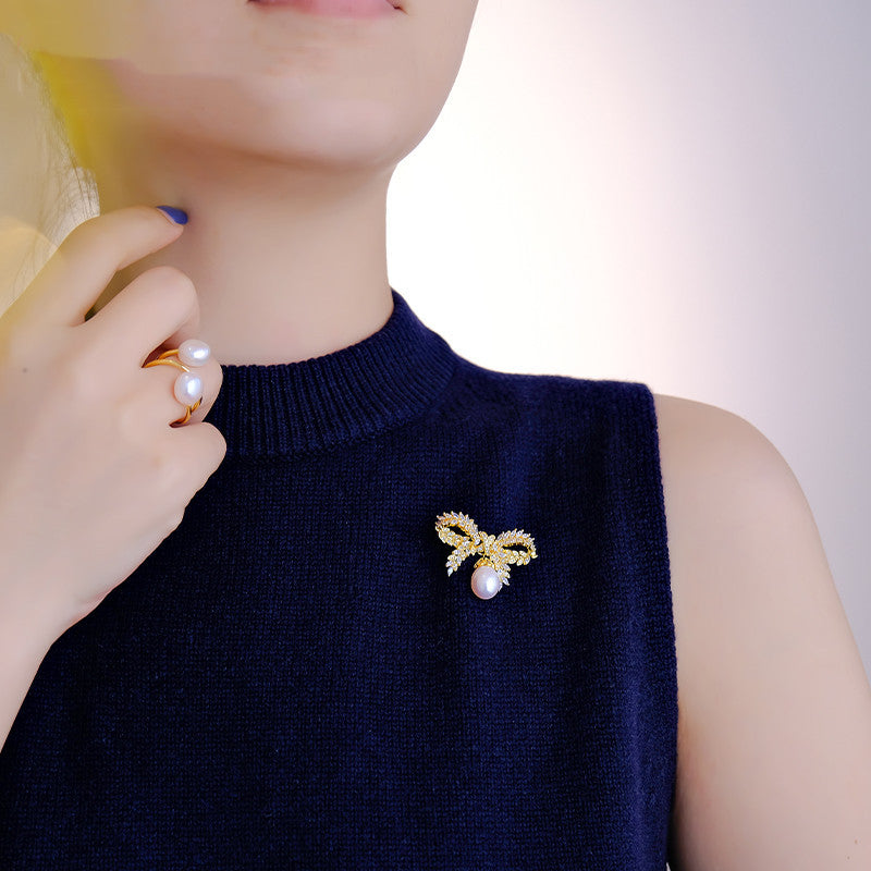 Person wearing a navy blue sleeveless top with a gold brooch and pearl detail, holding a pearl earring.