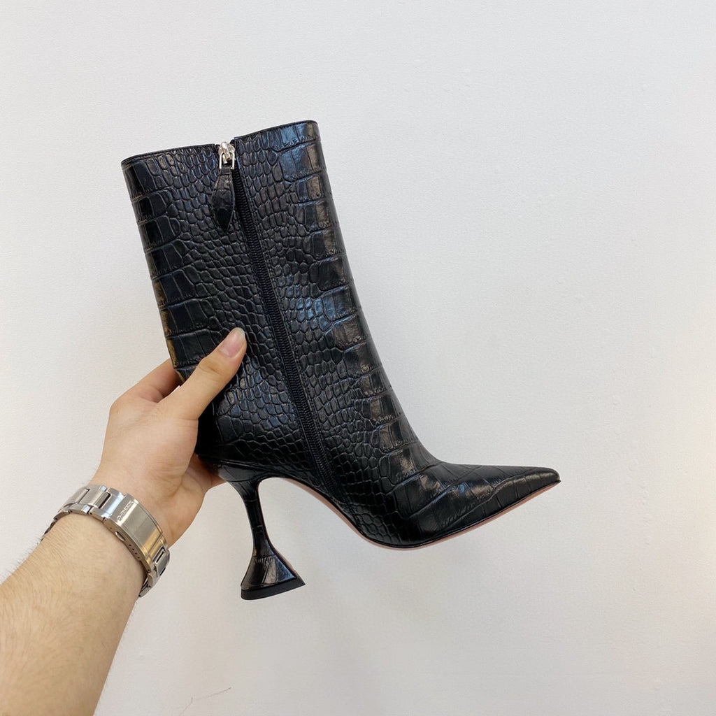 Black crocodile-patterned high-heeled boots held by a hand against a white background