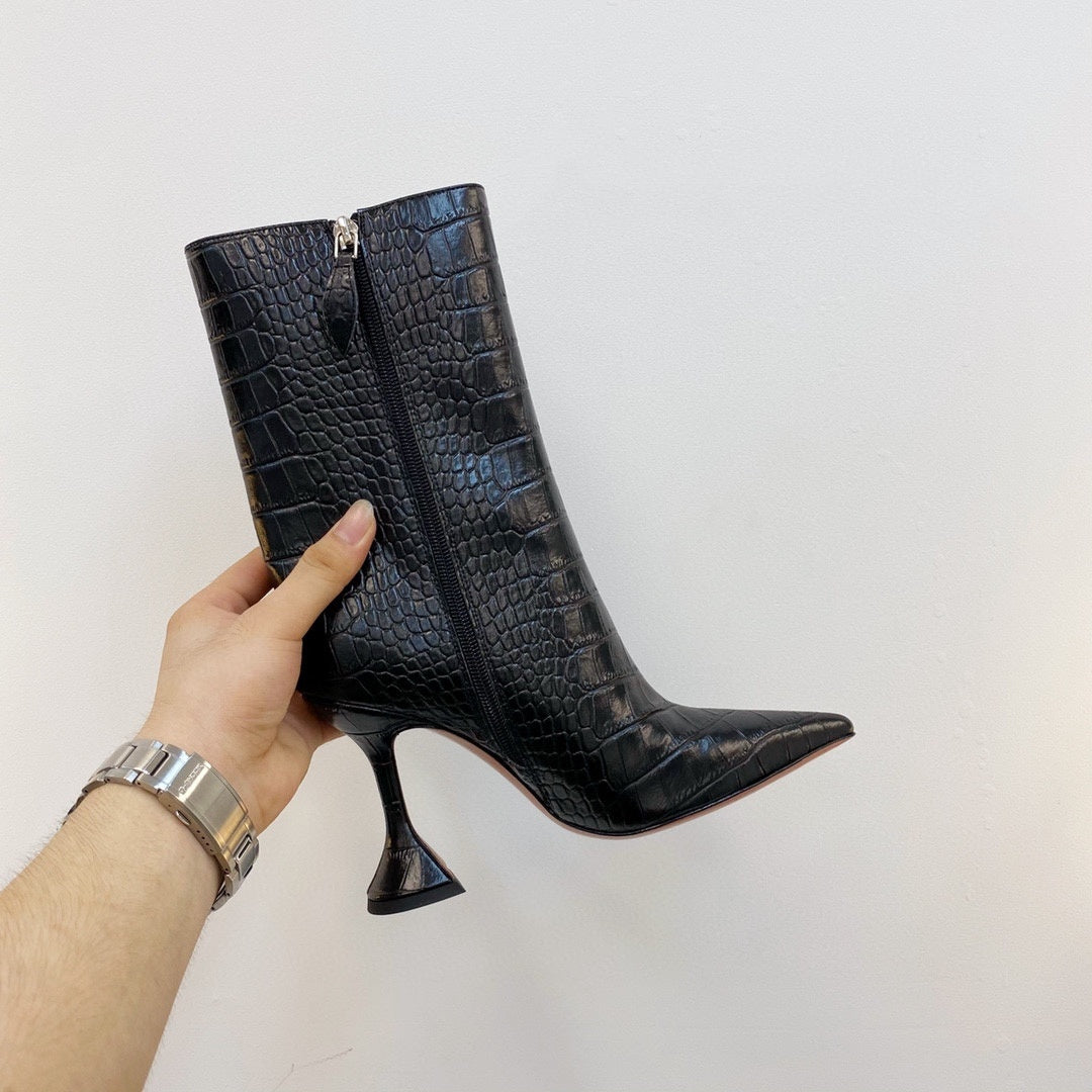 Black crocodile-patterned high-heeled boots held by a hand against a white background