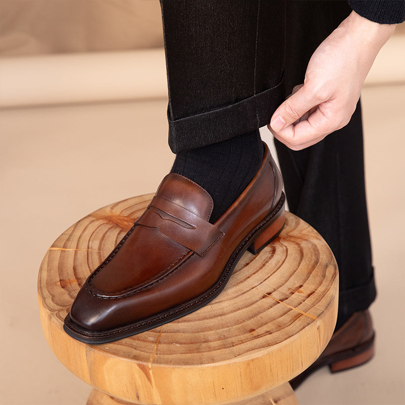Brown leather loafer being worn on a wooden stool with a neutral background