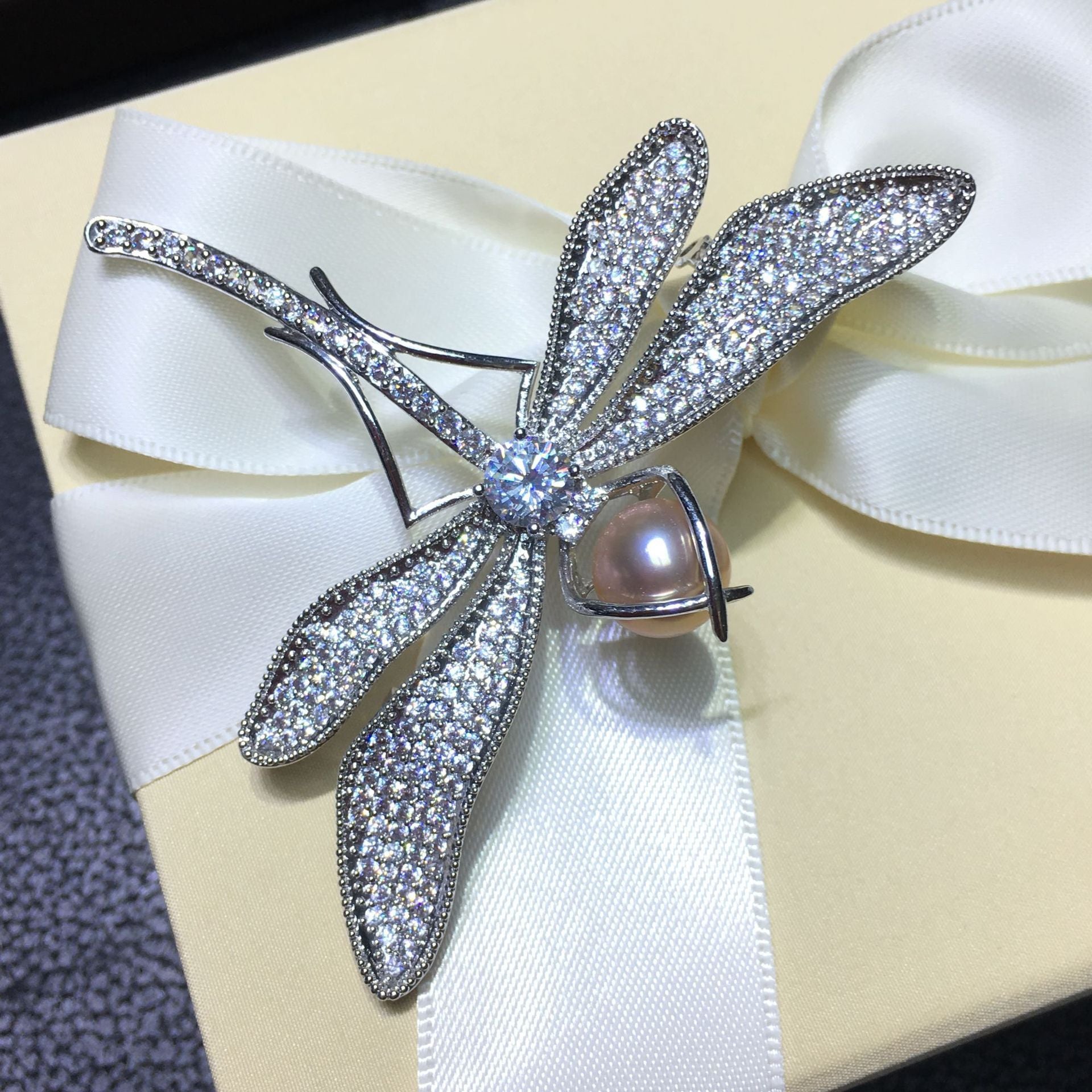 Silver dragonfly-shaped brooch with a pearl on a white ribbon