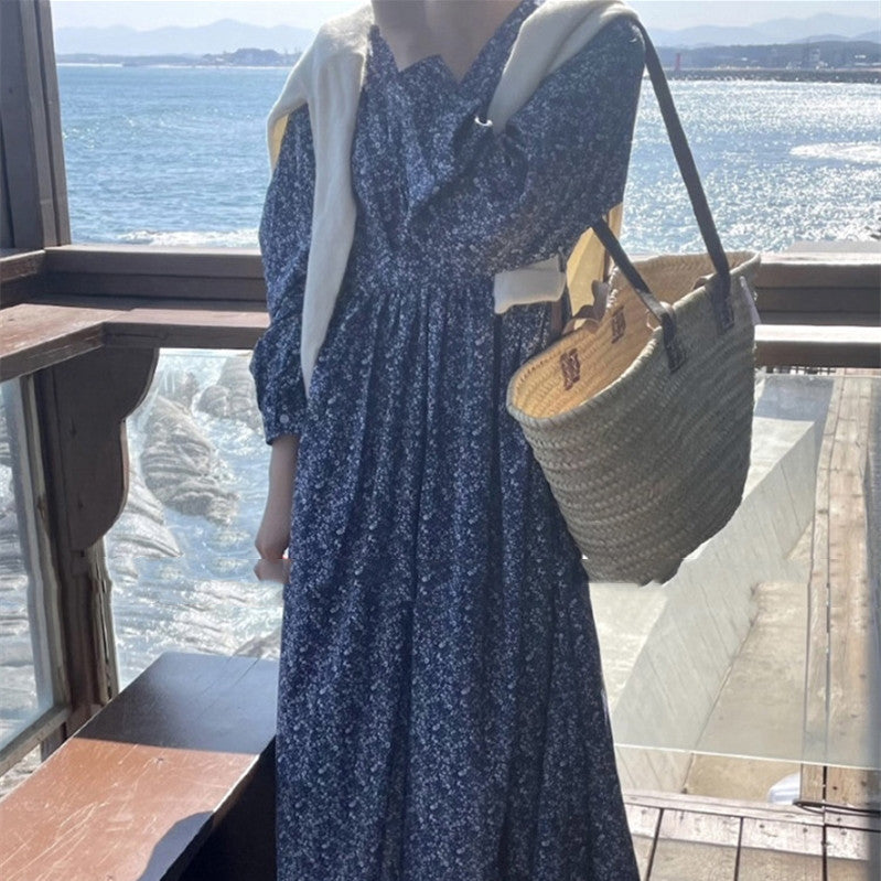 Person wearing a blue floral dress holding a straw bag by a waterfront.