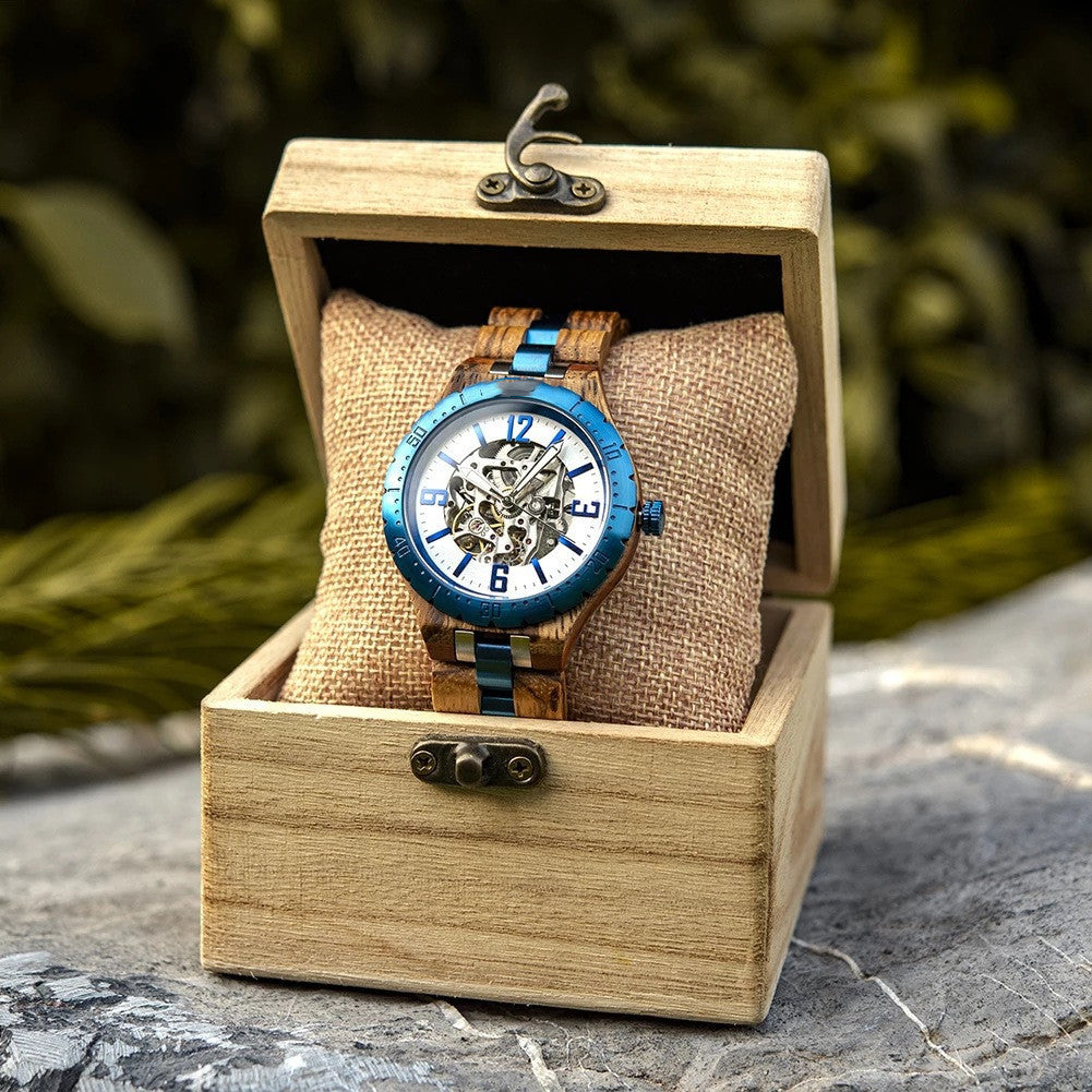 Wooden watch with blue face and brown strap in a wooden box on a stone surface with greenery in the background