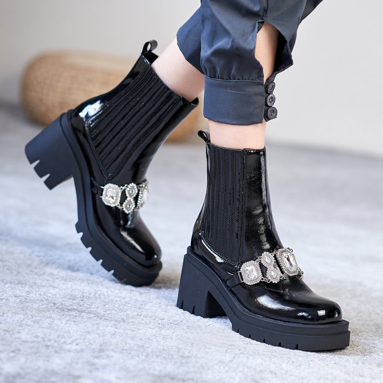 Black patent leather boots with decorative buckles worn by a person on a light gray surface.