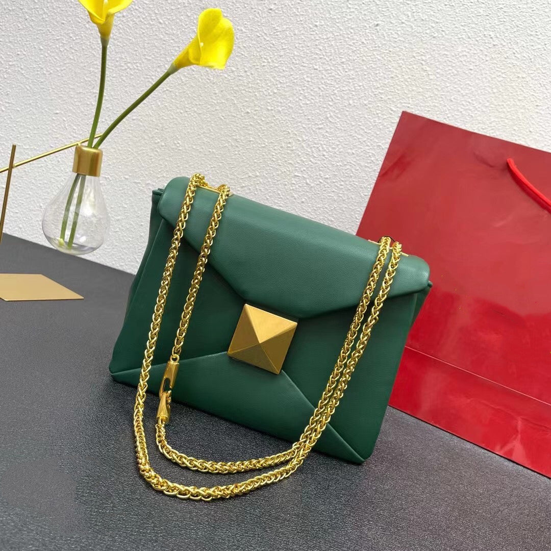Green handbag with gold chain and pyramid handle on a gray surface with yellow flowers and a red bag in the background.