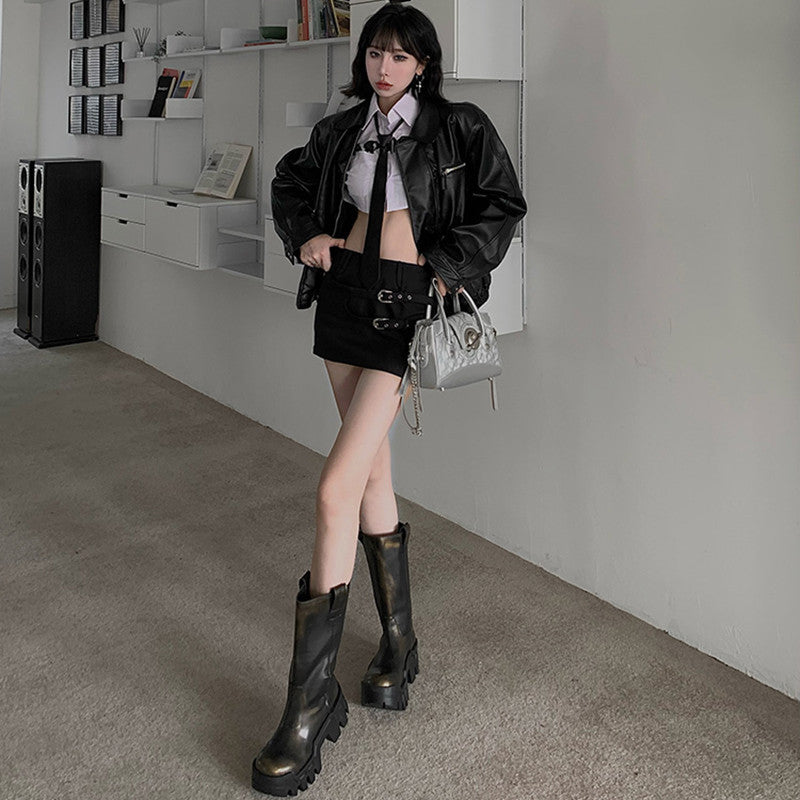 Person wearing a black leather jacket, white shirt, and black boots in an indoor setting.