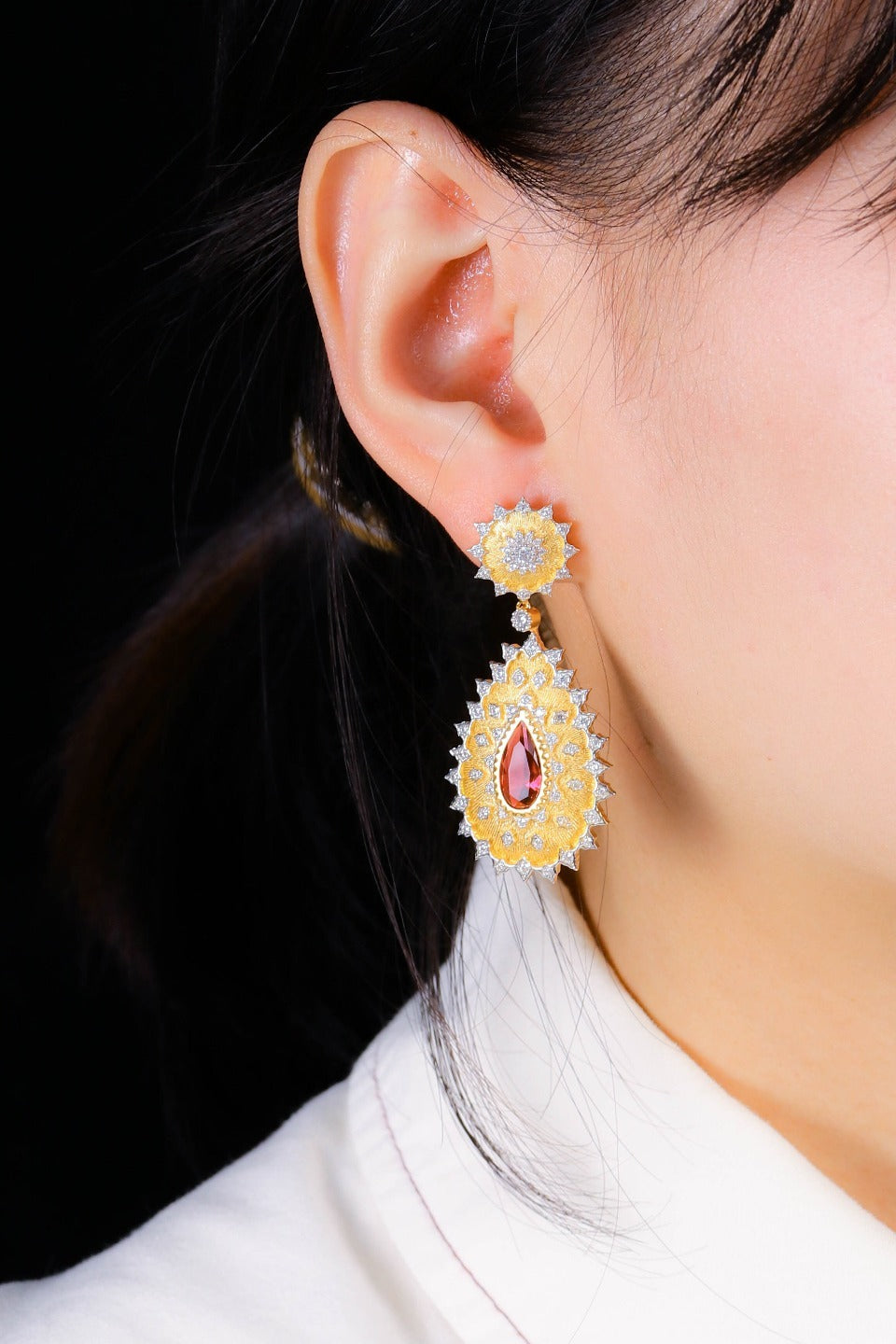 Gold earrings with a central red gemstone worn by a person against a dark background