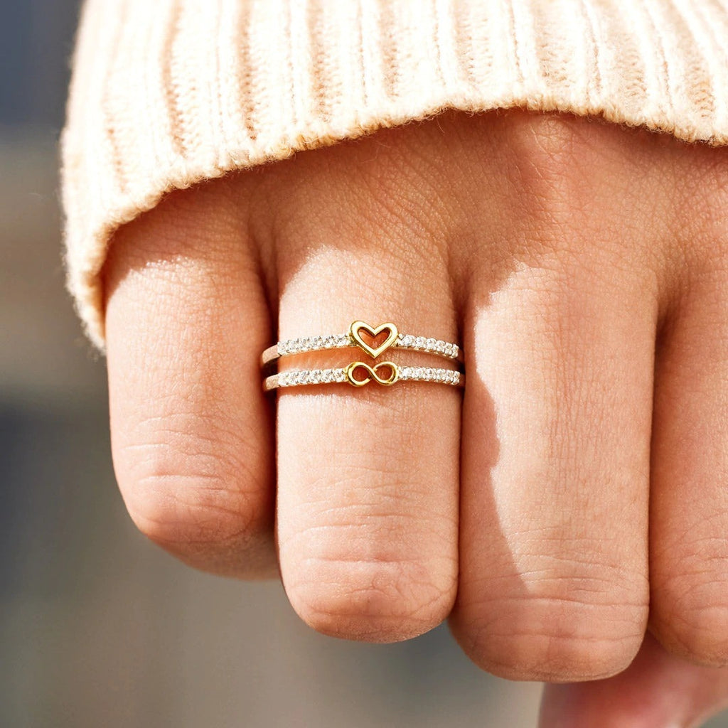 Close-up of a hand wearing two gold rings with heart and infinity symbols on a blurred background.