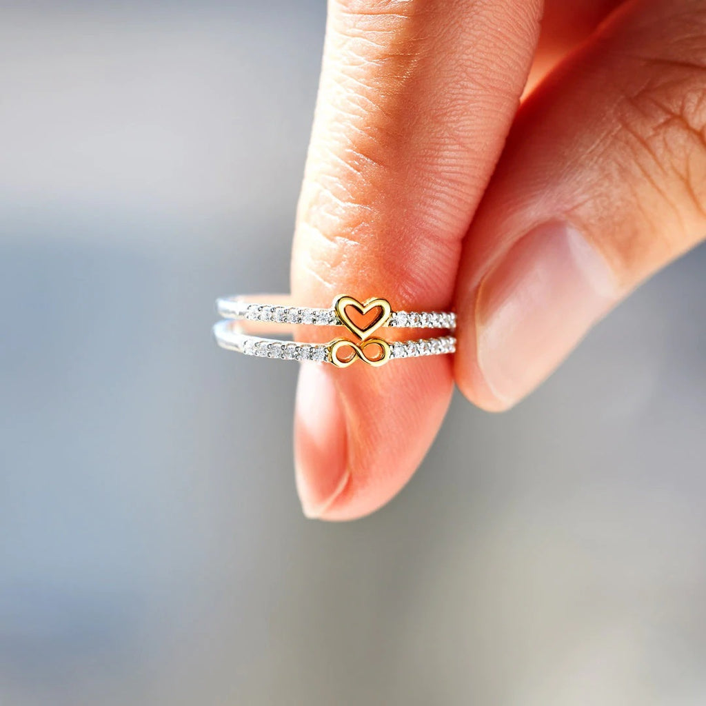 Close-up of a hand wearing two rings with heart and infinity symbol designs on a blurred background