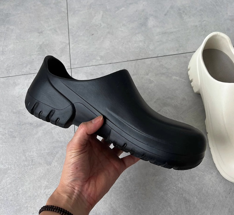 Black slip-on shoe held by a hand with a white shoe in the background on a gray tiled floor.