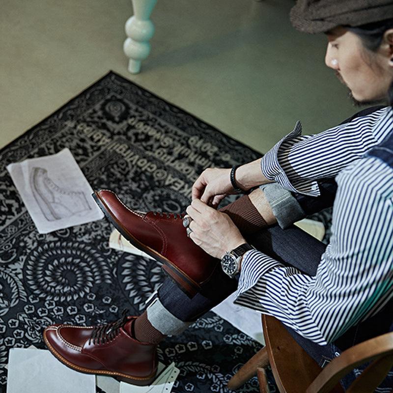 Person sitting on a chair wearing brown leather shoes, with a patterned rug and a book in the background.