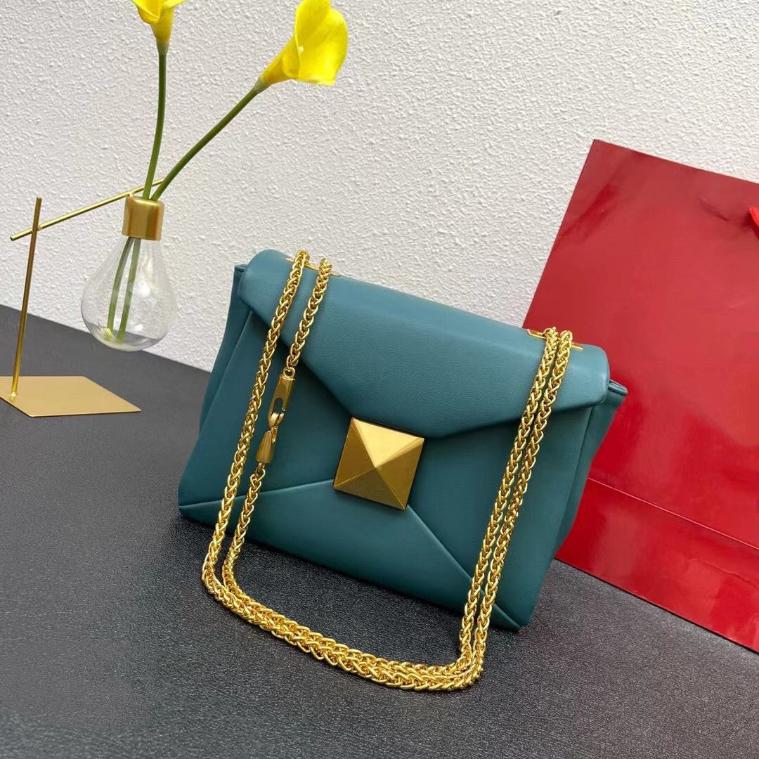 Teal handbag with gold chain and pyramid lock on a gray surface with a red bag and yellow flowers in the background.