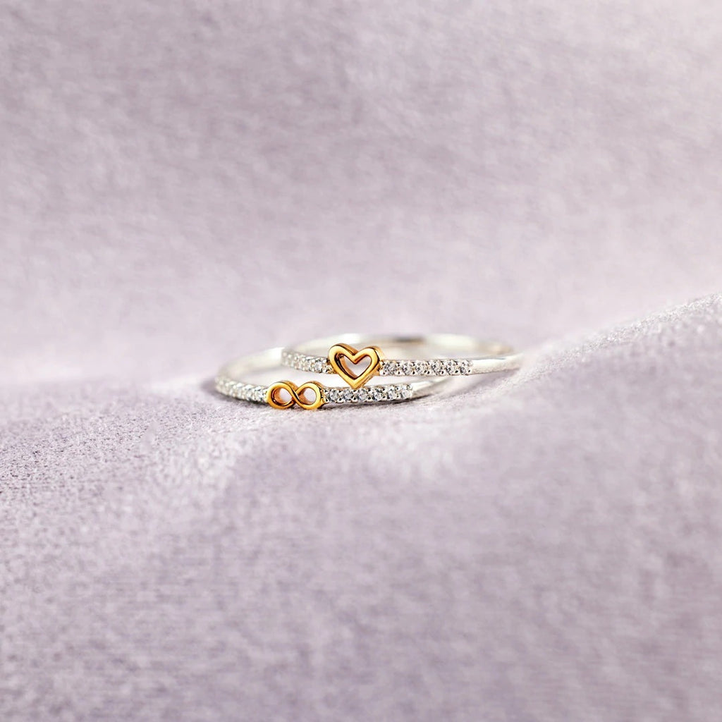 Two rings with heart and infinity symbol designs on a light purple fabric background