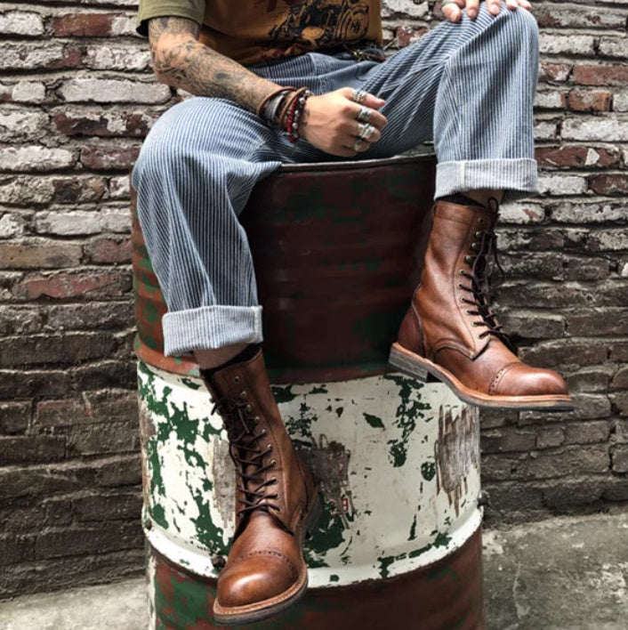Person wearing brown leather boots sitting on a rustic barrel against a brick wall.