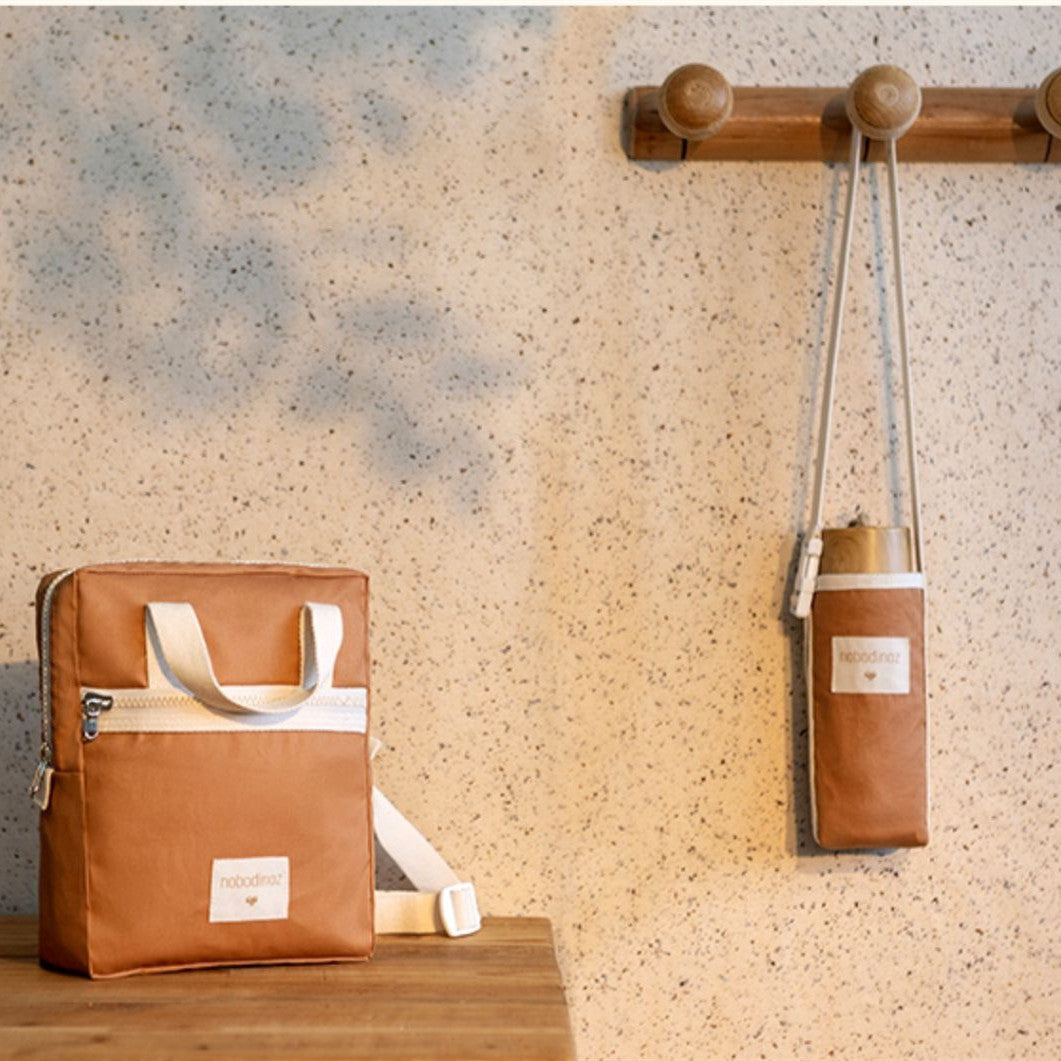 Brown leather bag with white strap on a wooden surface and wall-mounted hook with similar bag.