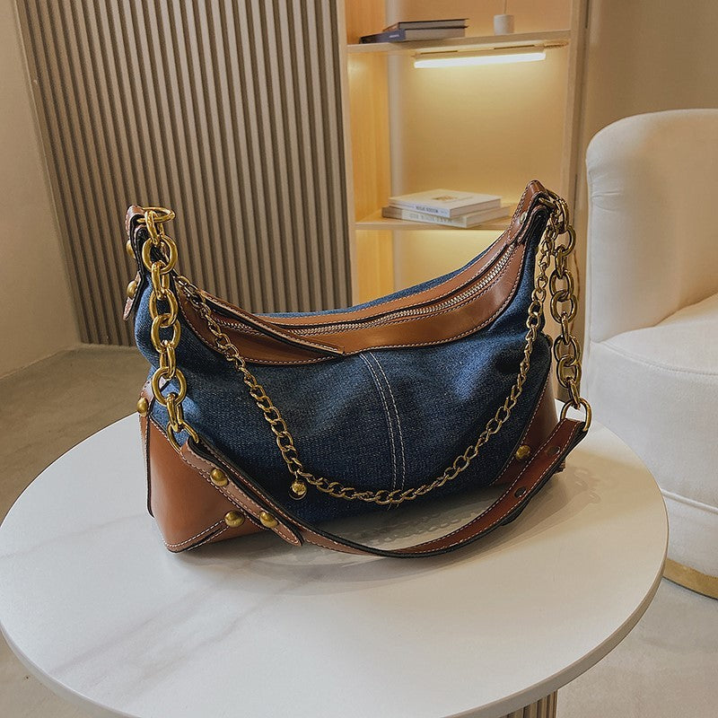 Denim handbag with brown leather accents and gold chain details on a white surface.