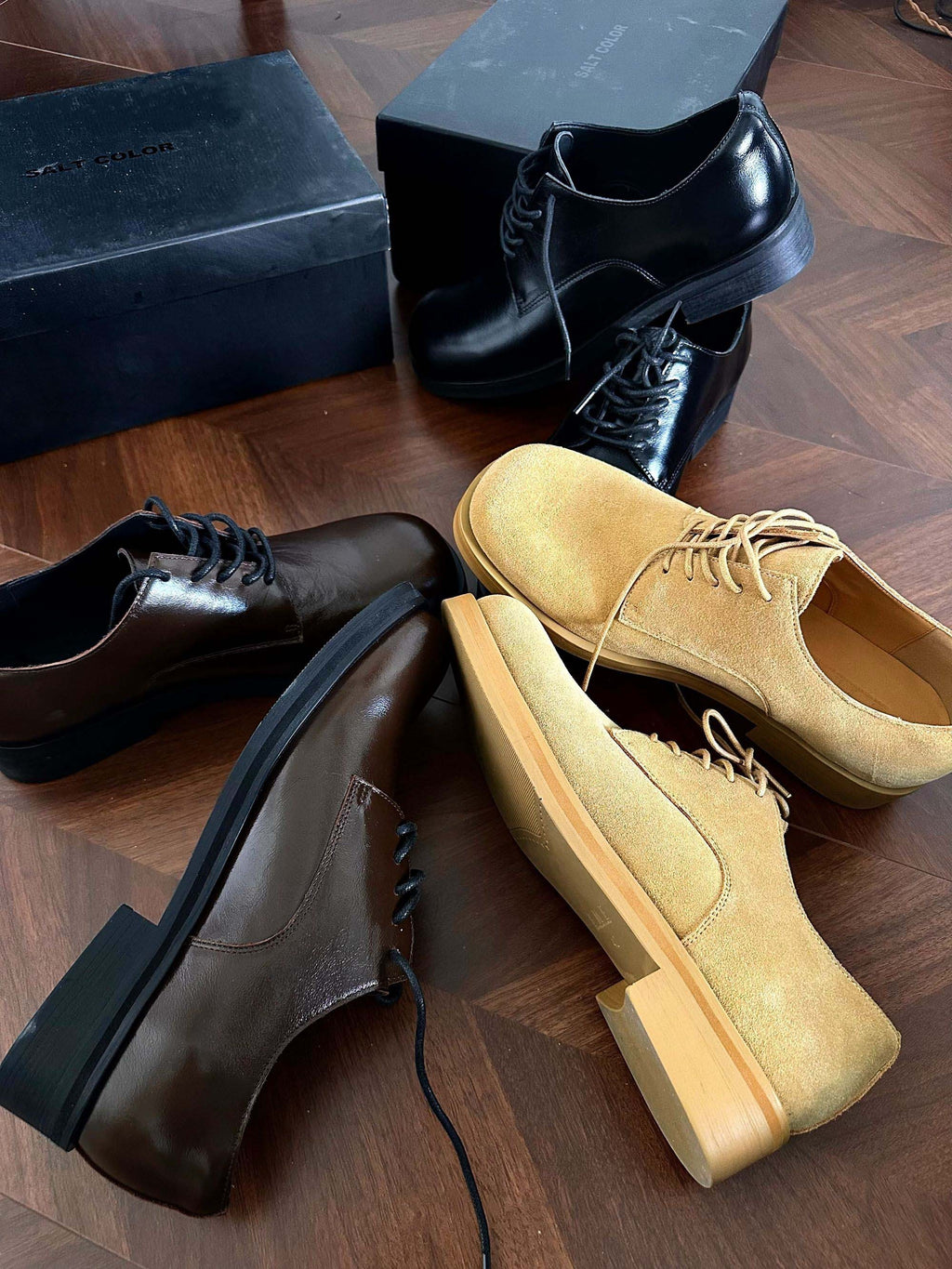 Three pairs of shoes including black, brown, and yellow on a wooden floor with a box in the background.