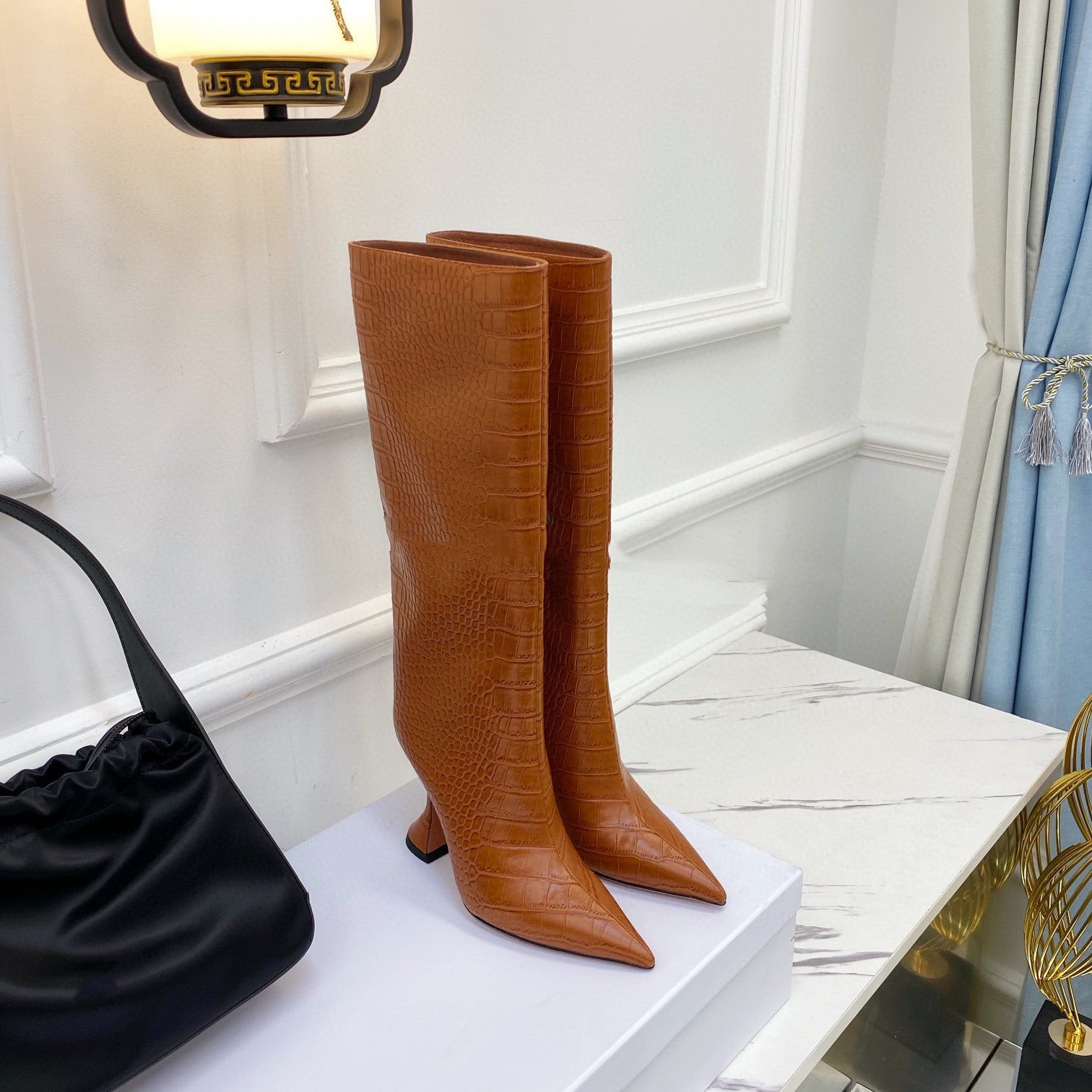 Brown knee-high boots on a white surface with a black handbag and decorative elements in the background.