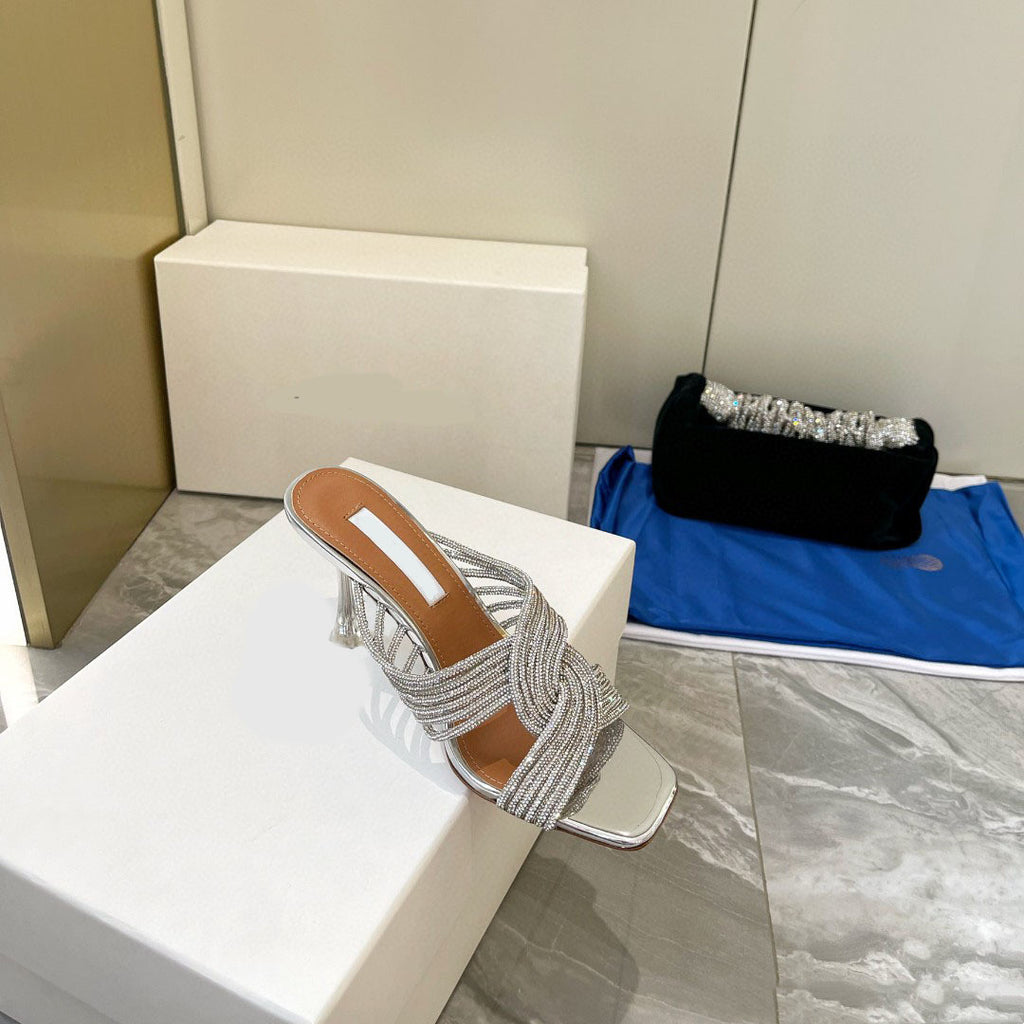 Silver sandal with fringe on a white block, with a black clutch and blue bag in the background.