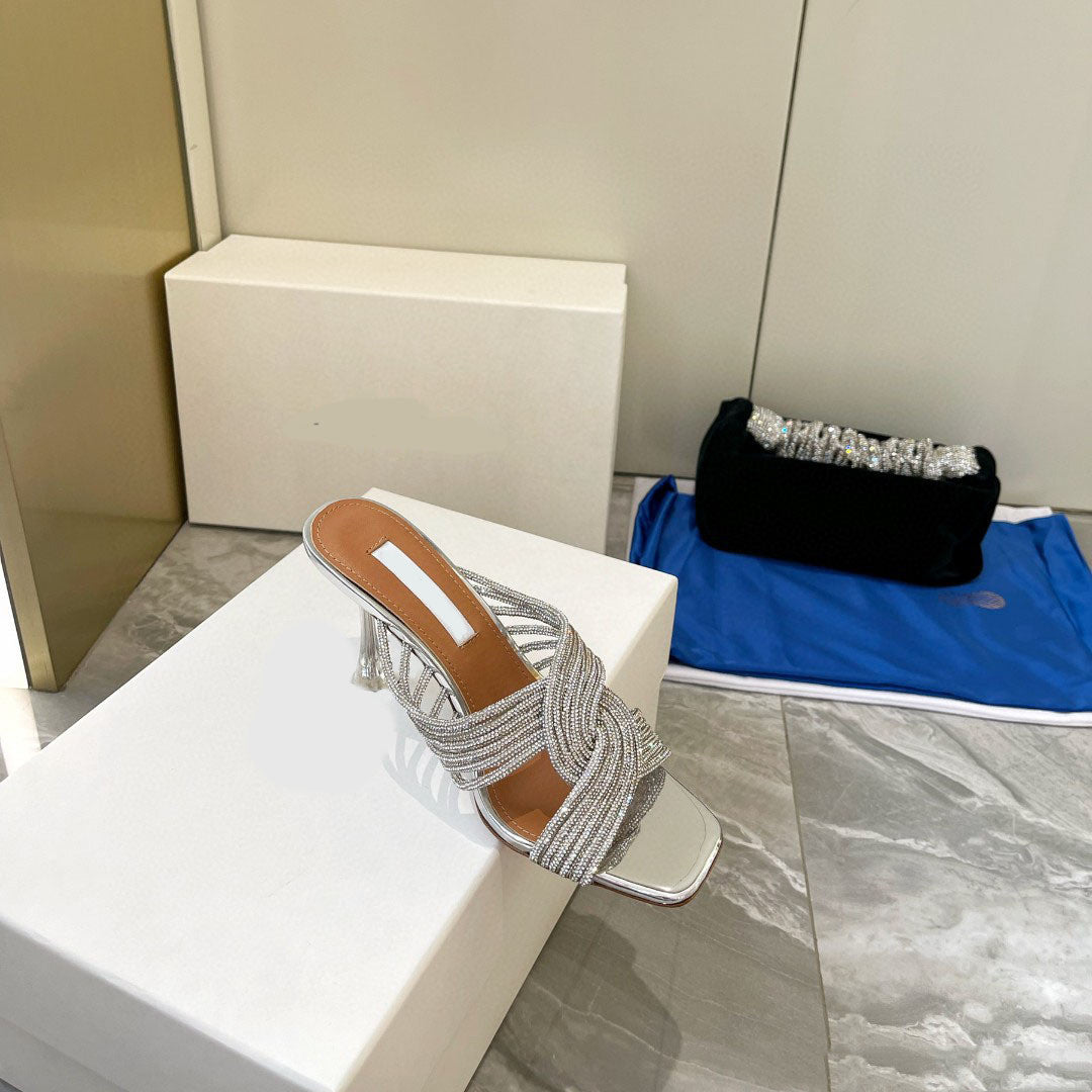 Silver sandal with fringe on a white block, with a black clutch and blue bag in the background.