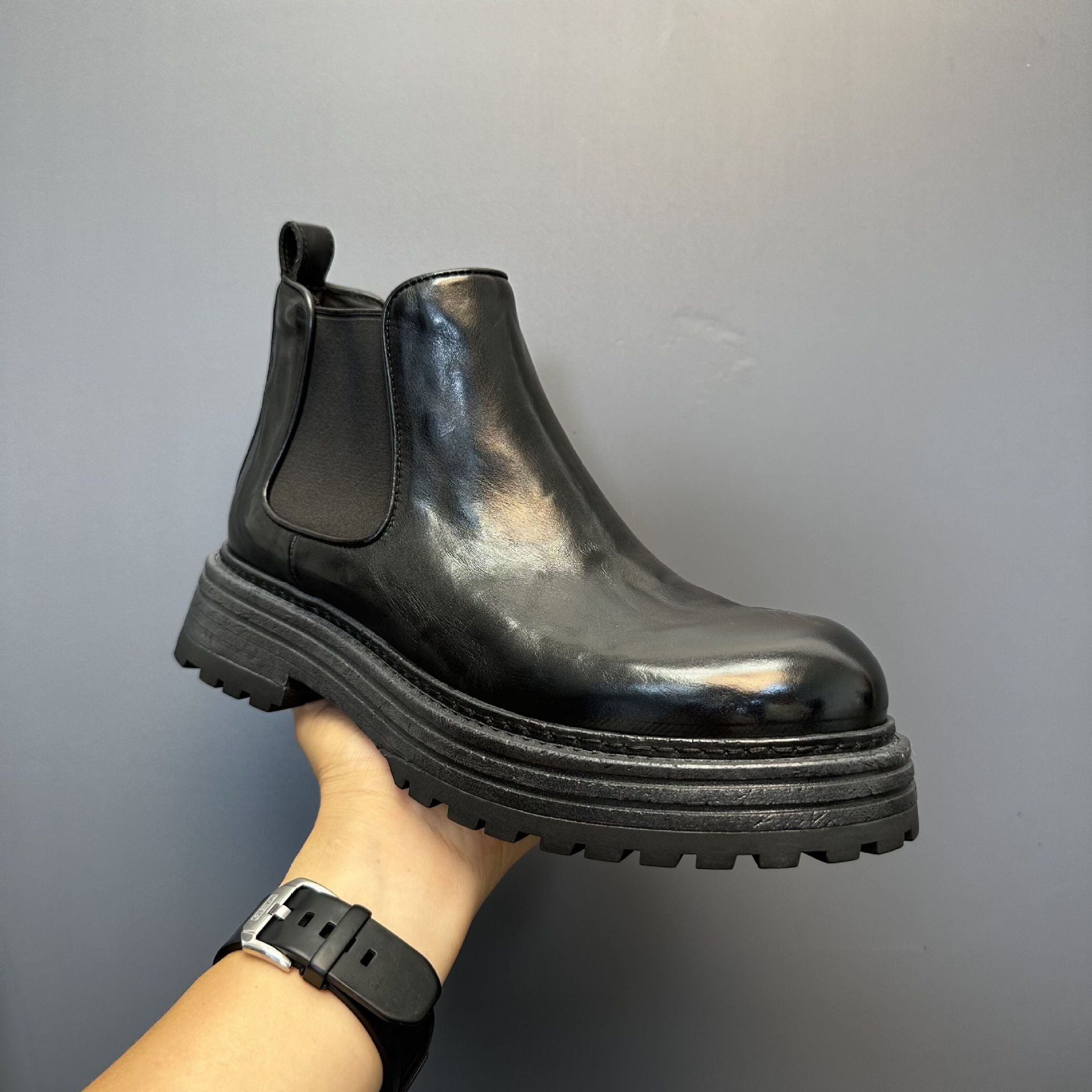 Black Chelsea boot held by a hand against a gray background