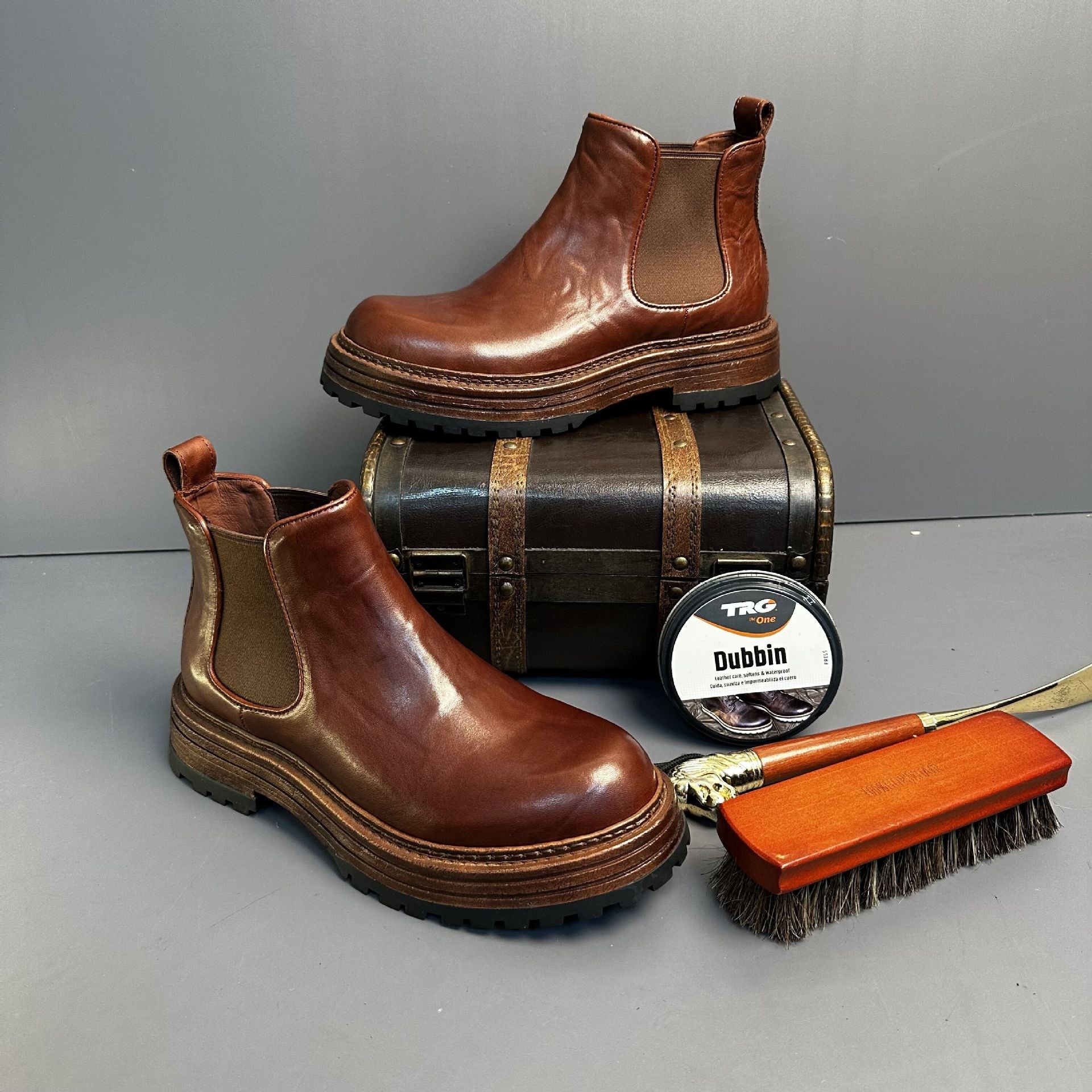 Brown leather boots with a barrel, TRG Dubbin container, and brush on a gray background