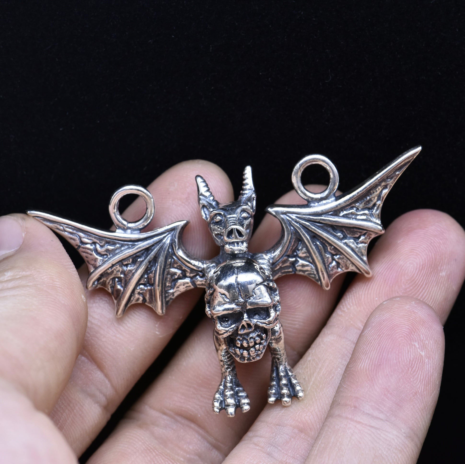 Silver bat-shaped pendant with a skull design held between fingers against a black background
