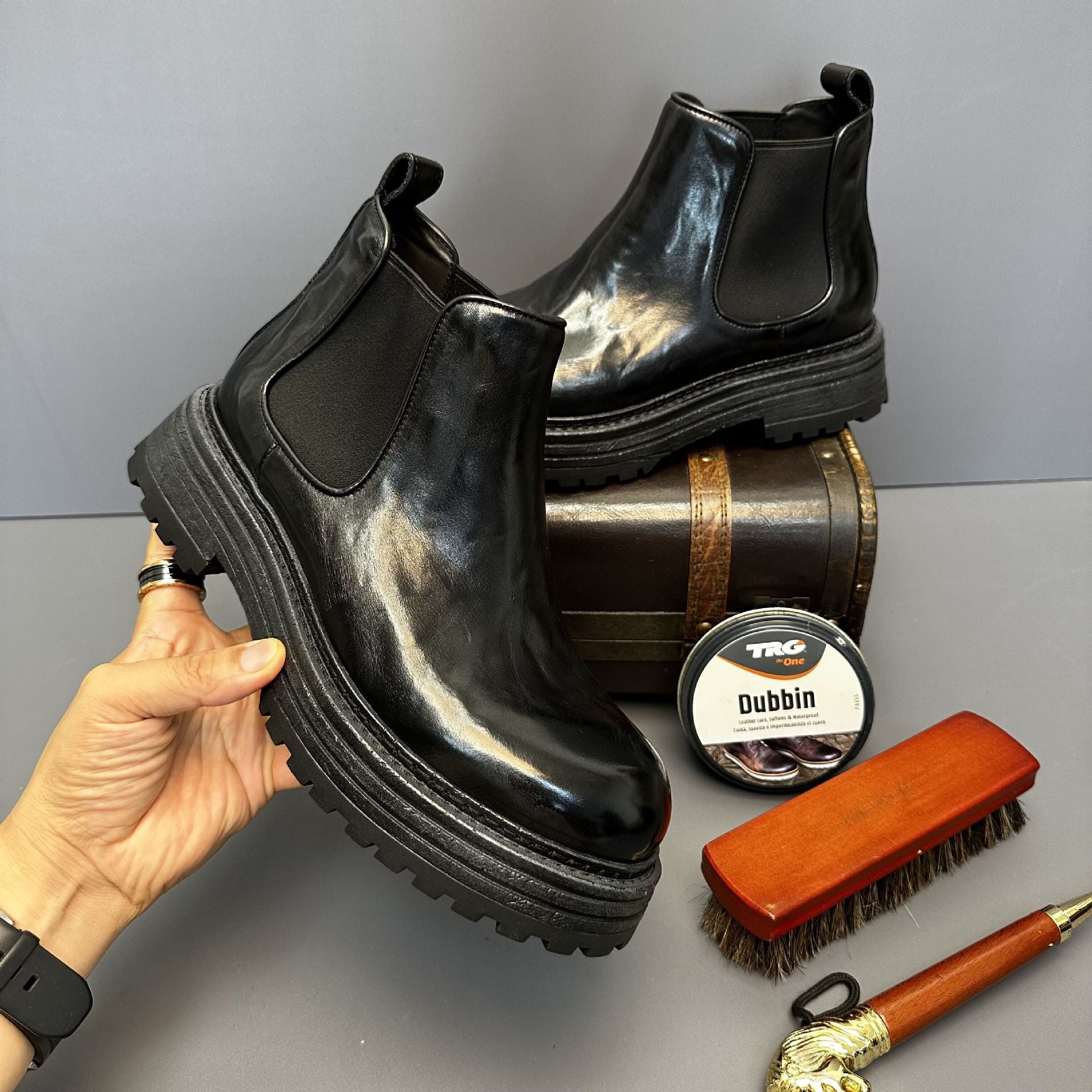 Black leather boot being held, with a barrel, TRG Dubbin container, and brush on a gray surface.