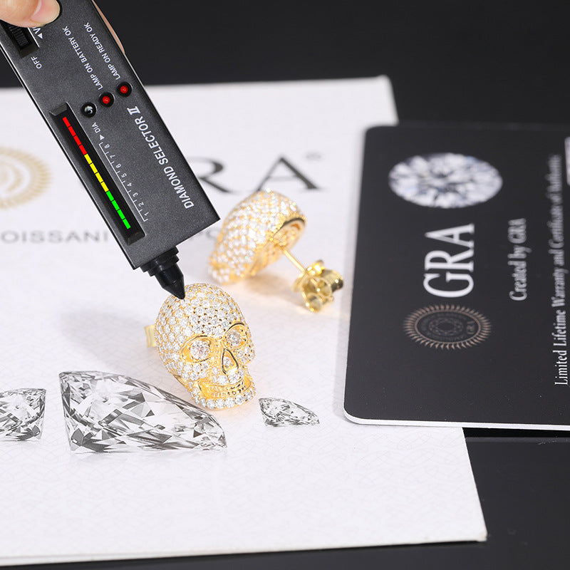 Gold skull-shaped earrings with diamonds on a white surface, next to a GRA card and diamond model.