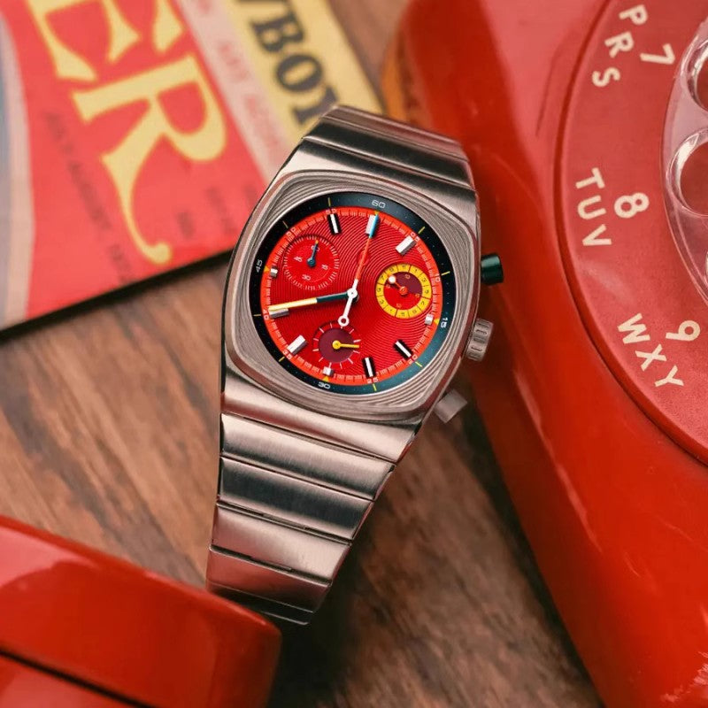 Watch with a red face and silver case on a wooden surface with a red rotary phone.