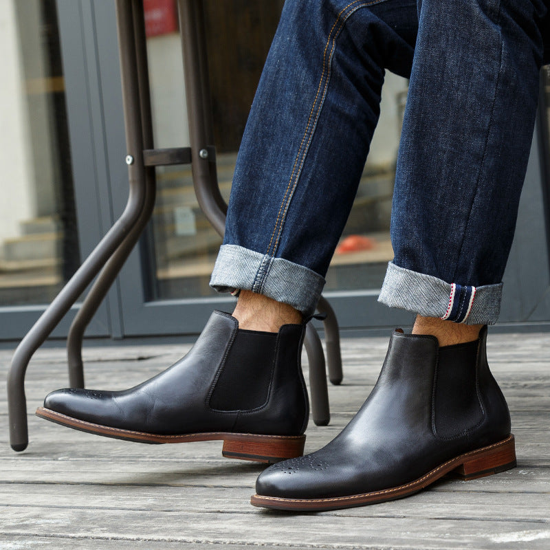 Person wearing black leather Chelsea boots with brown soles on a wooden floor.