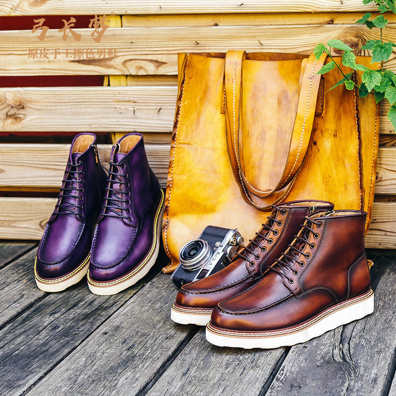 Pair of purple leather boots and a pair of brown leather boots on a wooden surface with a yellow bag and camera in the background.